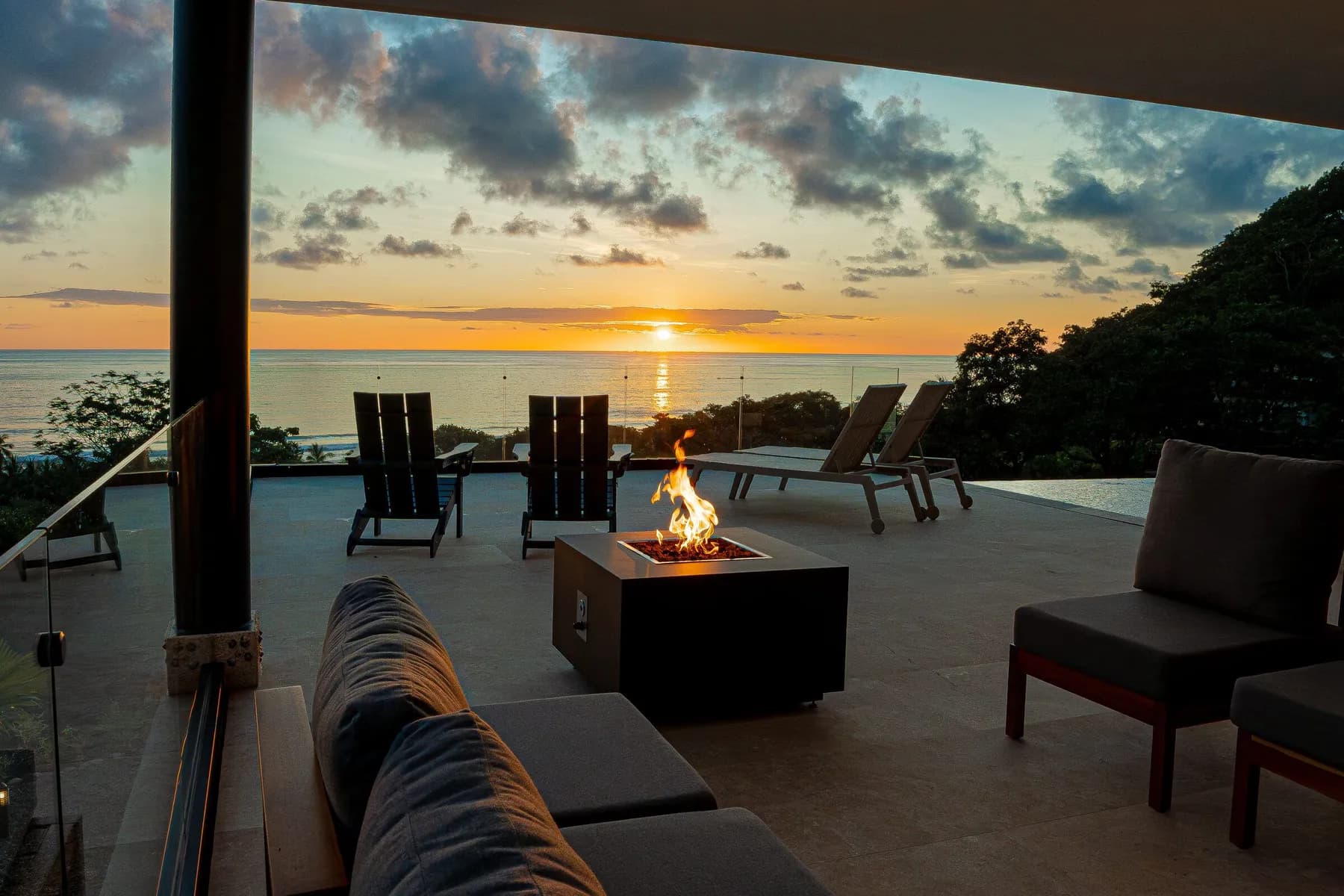 A lit firepit on a terrace at sunset, dark chairs facing the ocean and an orange glow on the horizon.
